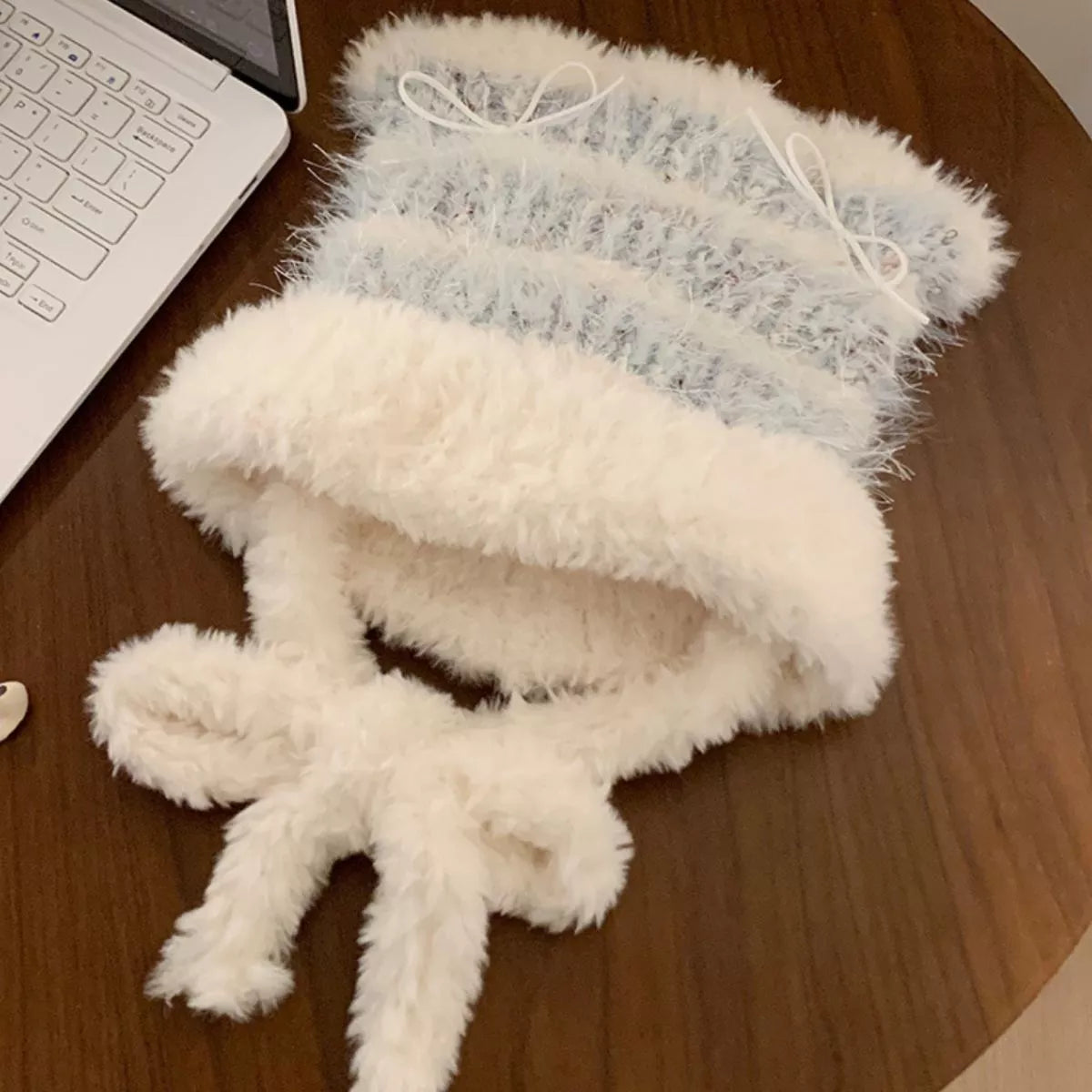 Fluffy white and gray knitted hat on a wooden surface with a laptop.