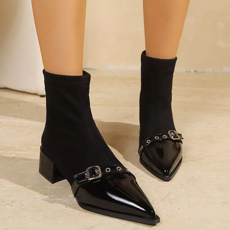 Black ankle boots with a glossy patent leather pointed toe, low block heel, and silver buckle strap details, shown on a model's feet against a neutral beige background.