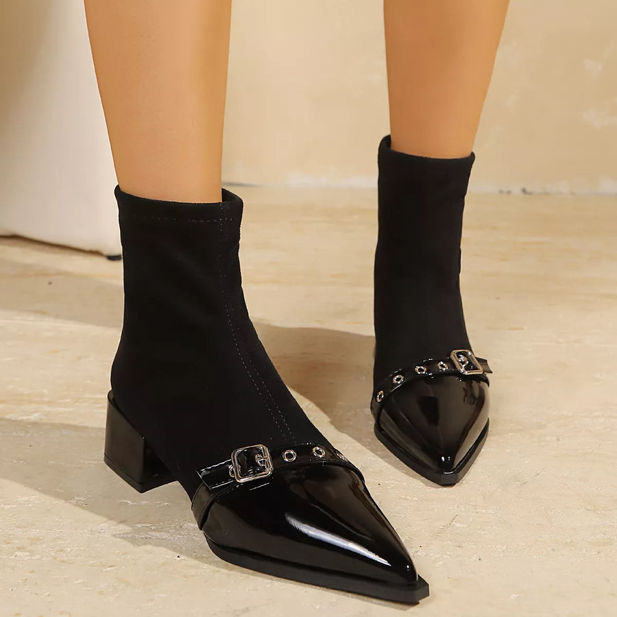 Black ankle boots with a glossy patent leather pointed toe, low block heel, and silver buckle strap details, shown on a model's feet against a neutral beige background.