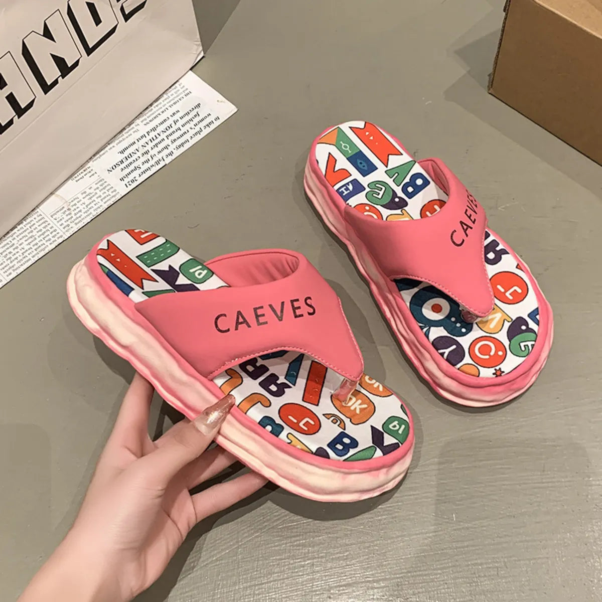 Pink slippers with colorful letter pattern held by a hand on a gray surface.