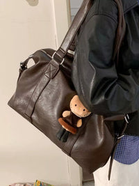 Brown leather duffel bag with a small plush toy inside, worn by a person in a black jacket.