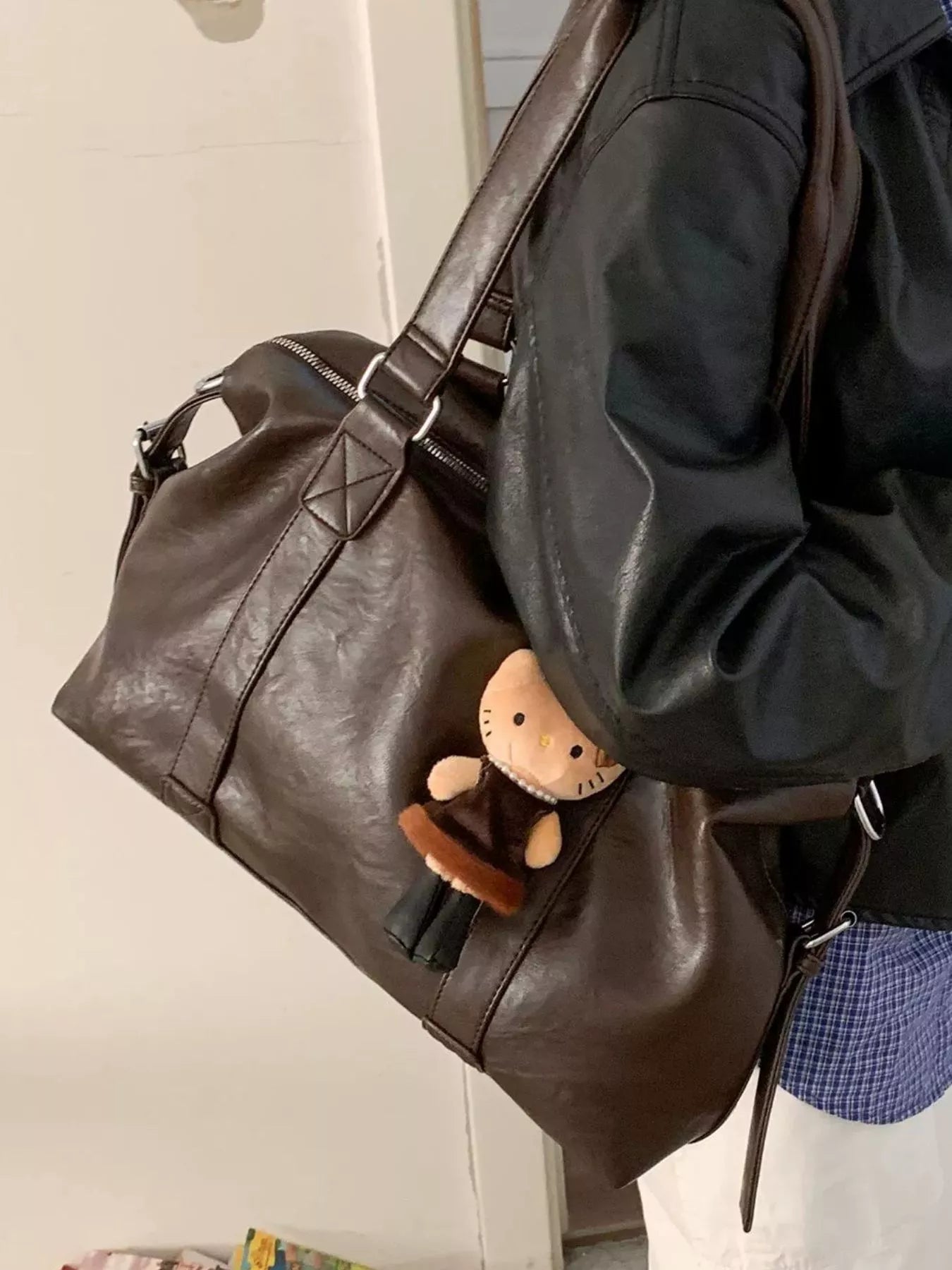 Brown leather duffel bag with a small plush toy inside, worn by a person in a black jacket.