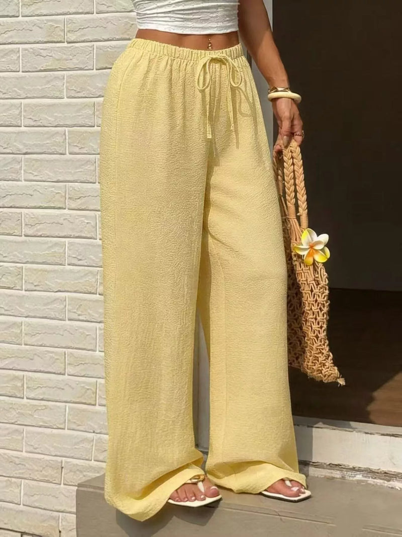 Person wearing yellow wide-leg pants holding a woven bag against a stone wall.