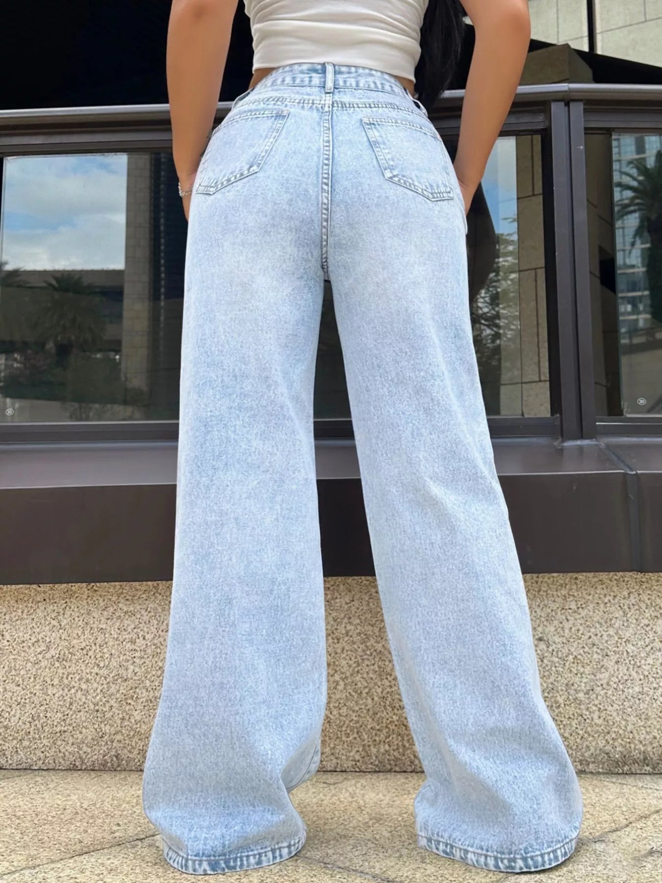 Person wearing light blue jeans standing in front of a building with reflective glass windows.