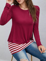 A woman wearing a long-sleeve crewneck top in deep burgundy, paired with a red and white striped shirt tucked into blue jeans.