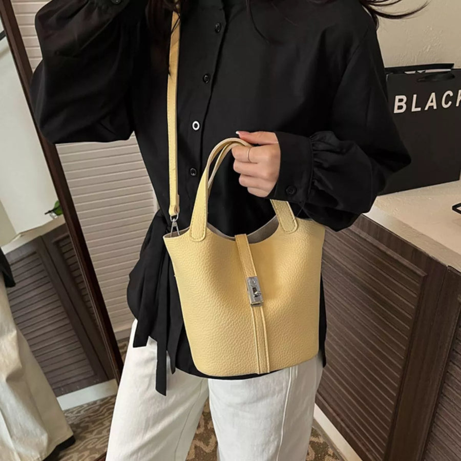 A person wearing a black button-up shirt and white pants carries a light yellow pebbled leather bucket tote bag with a top handle and shoulder strap in an indoor setting.