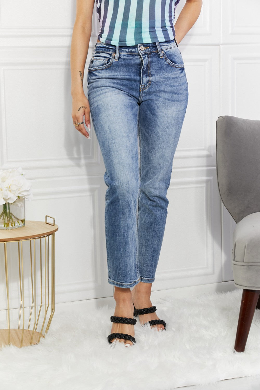 Person wearing blue jeans and a striped top in a room with a white wall and gray chair.