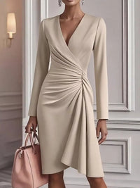 Woman wearing a beige long-sleeve dress holding a pink handbag in an elegant interior setting.