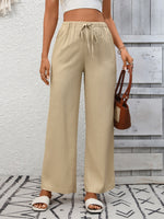 Person wearing beige wide-leg pants with a white top and sandals, holding a brown handbag against a light gray wall.