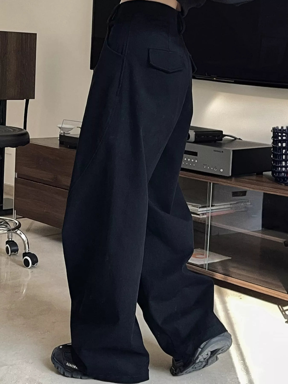 A person wearing loose, wide-leg black pants and black sneakers, standing in a modern living room with a TV and media console in the background.