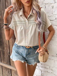 Woman wearing a beige button-up shirt and denim shorts, holding a straw bag against a white brick wall.