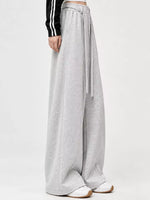 A person wearing light gray wide-leg sweatpants with a drawstring waist, paired with white sneakers and a black long-sleeve top with white stripes, shown against a plain light gray studio background.