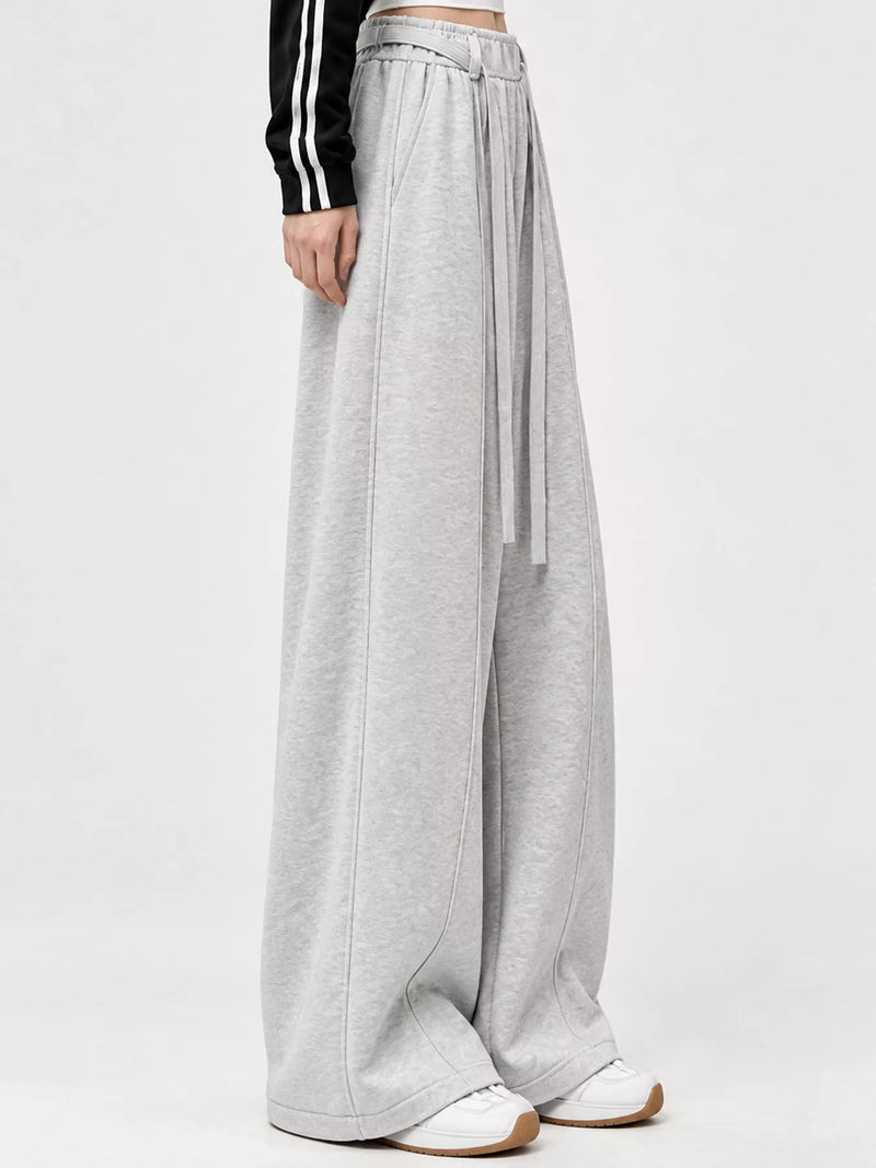 A person wearing light gray wide-leg sweatpants with a drawstring waist, paired with white sneakers and a black long-sleeve top with white stripes, shown against a plain light gray studio background.