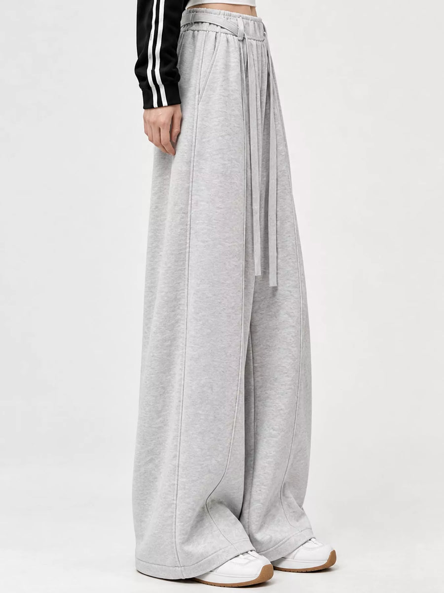 A person wearing light gray wide-leg sweatpants with a drawstring waist, paired with white sneakers and a black long-sleeve top with white stripes, shown against a plain light gray studio background.