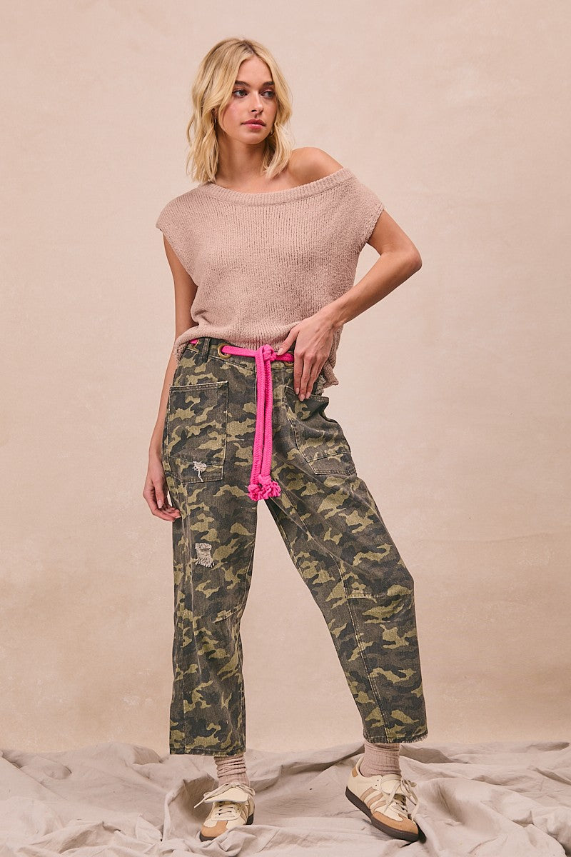 Woman wearing a pink top and camouflage pants with a pink belt against a beige background