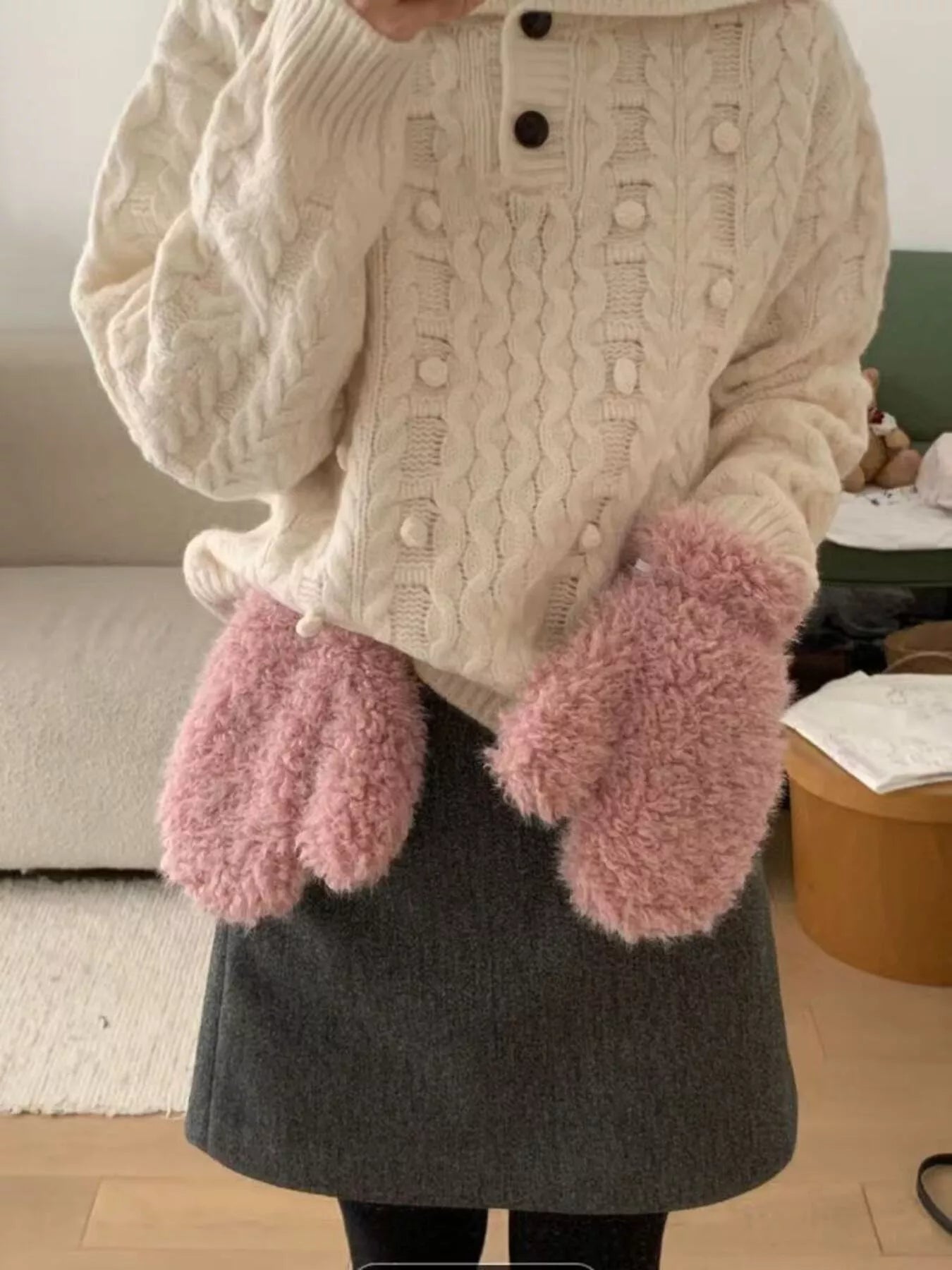 Person wearing a cream cable knit sweater and pink fuzzy mittens indoors.