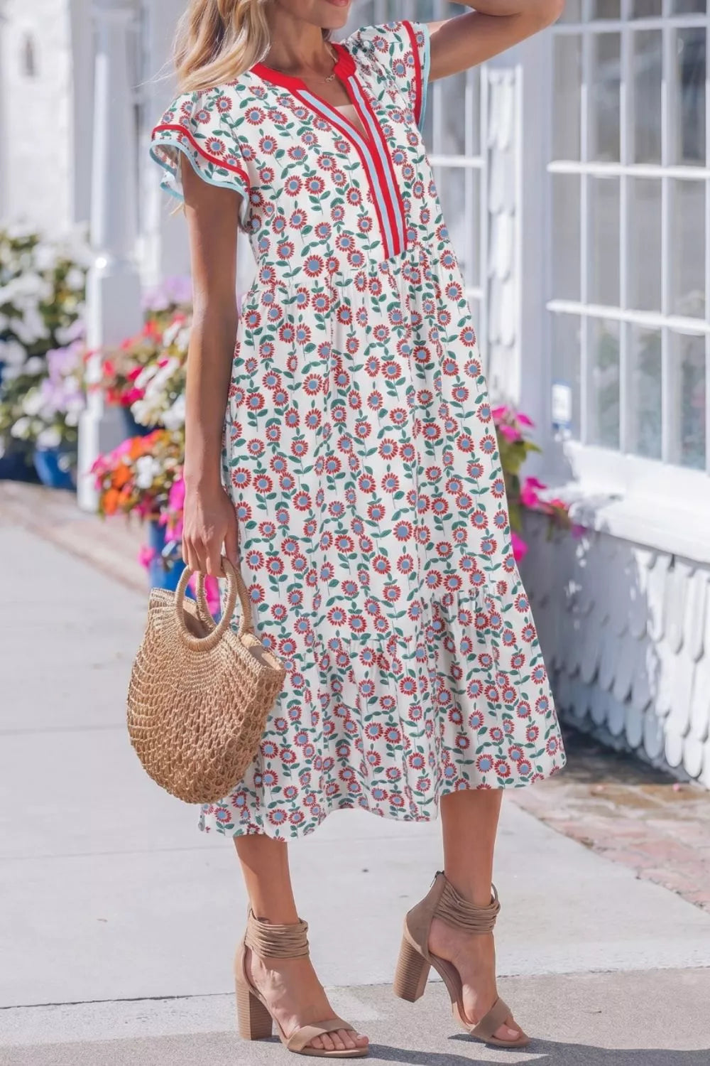Woman wearing a floral dress with red accents, holding a straw bag, standing on a porch.