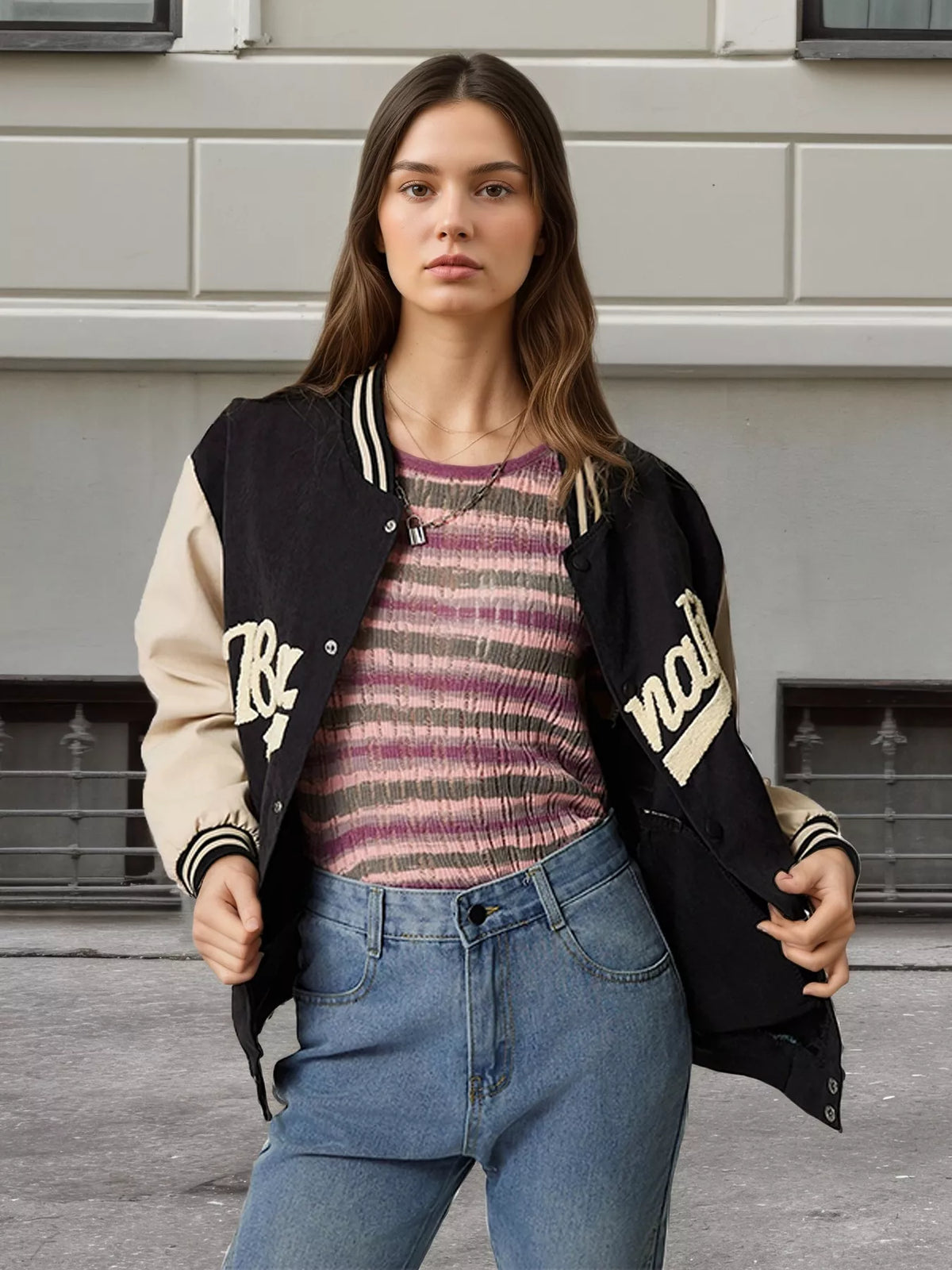 A woman wearing a varsity-style jacket with black and beige sleeves, layered over a striped knit top and blue jeans, standing outdoors against a light gray building wall.