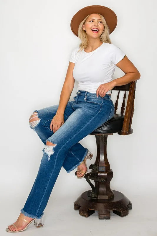 Woman sitting on a wooden chair wearing a white t-shirt, blue jeans, and a brown hat.
