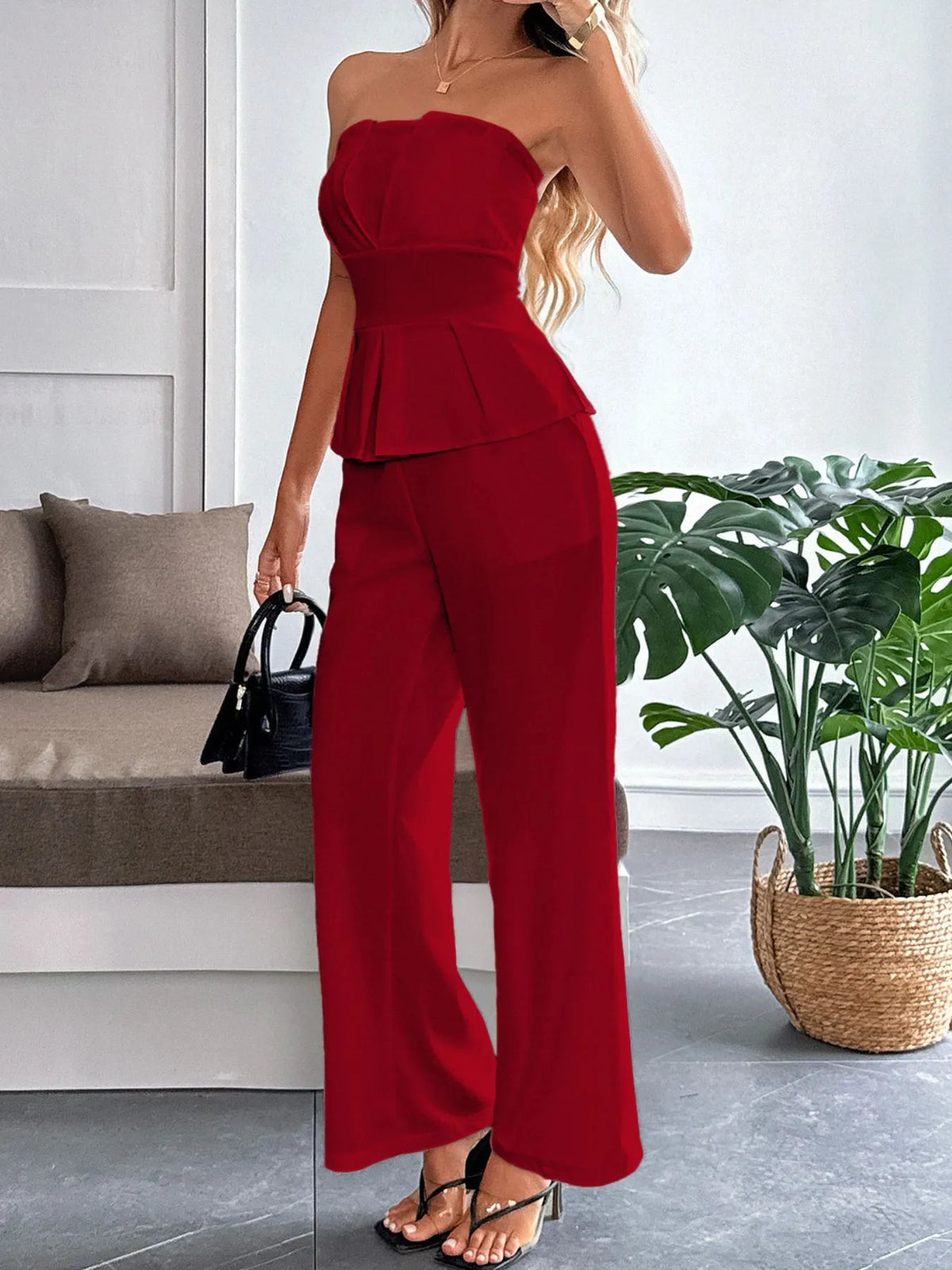 Woman in a red strapless top and wide-leg pants standing in a room with a plant and sofa.