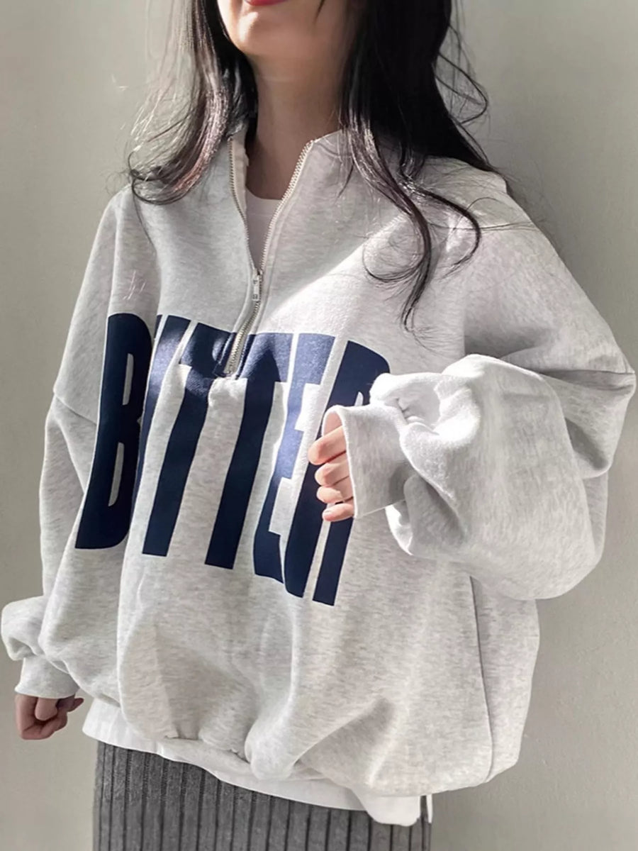 A person wearing an oversized light gray quarter-zip sweatshirt with large navy blue block letters spelling 'BITTER' across the front, paired with a gray ribbed skirt.