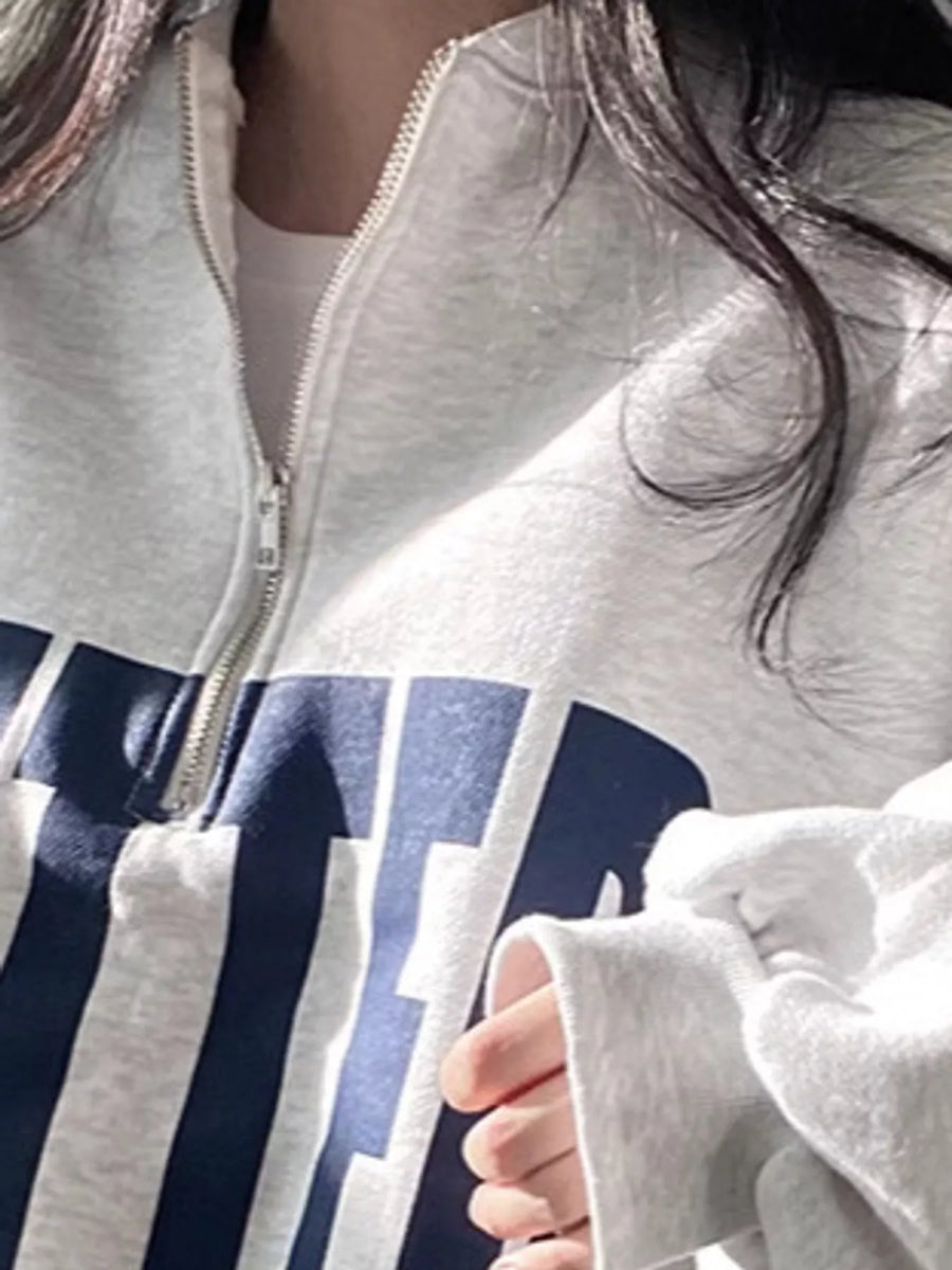 Close-up of a person wearing a light gray zip-up hoodie with a large navy blue graphic print on the front, paired with a white undershirt.