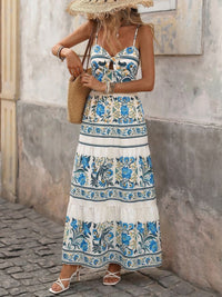 Woman wearing a floral dress with a straw hat and beige bag against a stone wall.