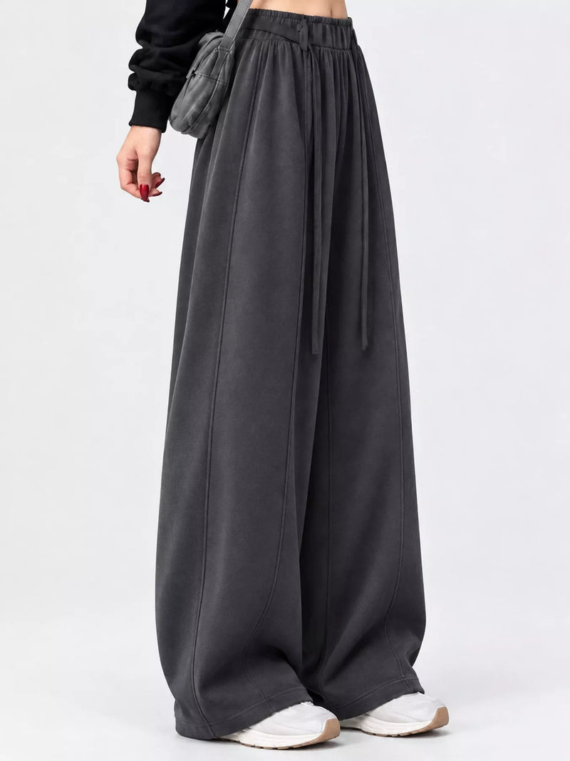 A person wearing very wide-leg dark gray lounge pants with an elastic waistband and drawstring, paired with white sneakers and a black top, photographed against a light gray studio background.