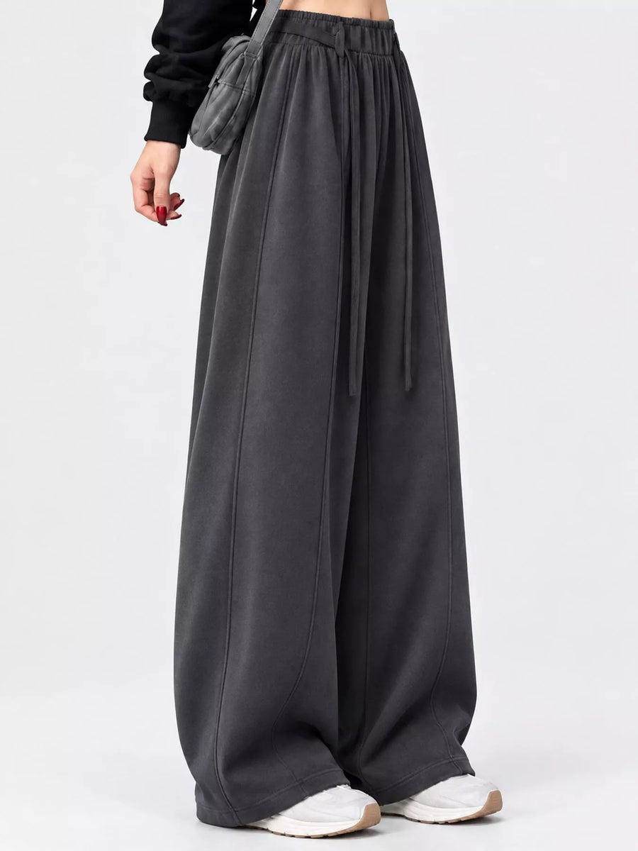 A person wearing very wide-leg dark gray lounge pants with an elastic waistband and drawstring, paired with white sneakers and a black top, photographed against a light gray studio background.