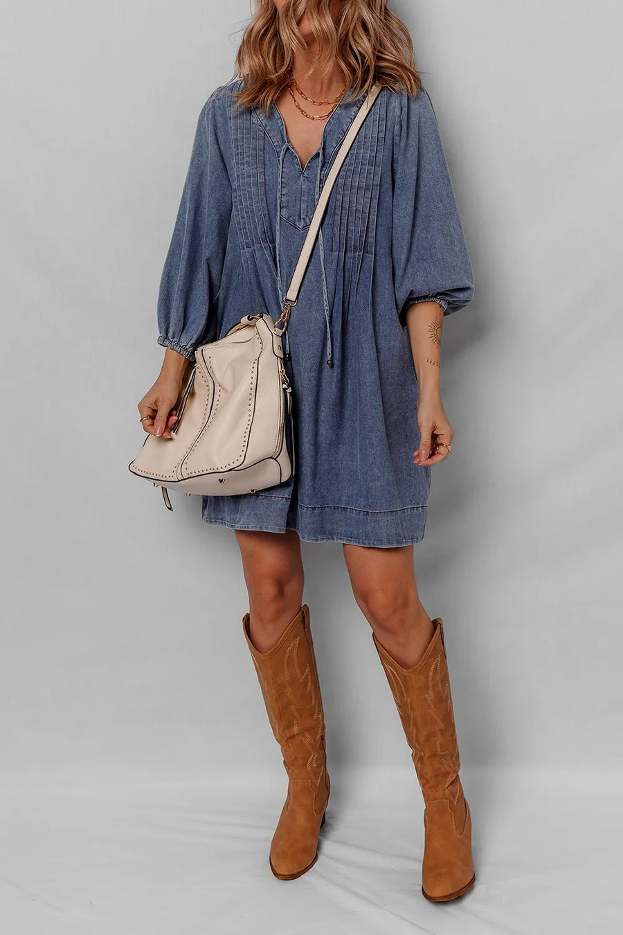 Person wearing a blue dress and brown boots holding a beige backpack on a plain background