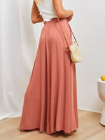 Person wearing a long pink skirt with a white top and straw bag against a plain background