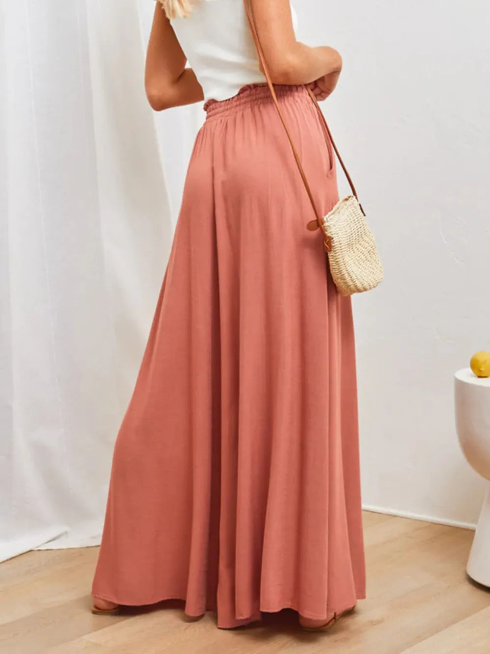 Person wearing a long pink skirt with a white top and straw bag against a plain background