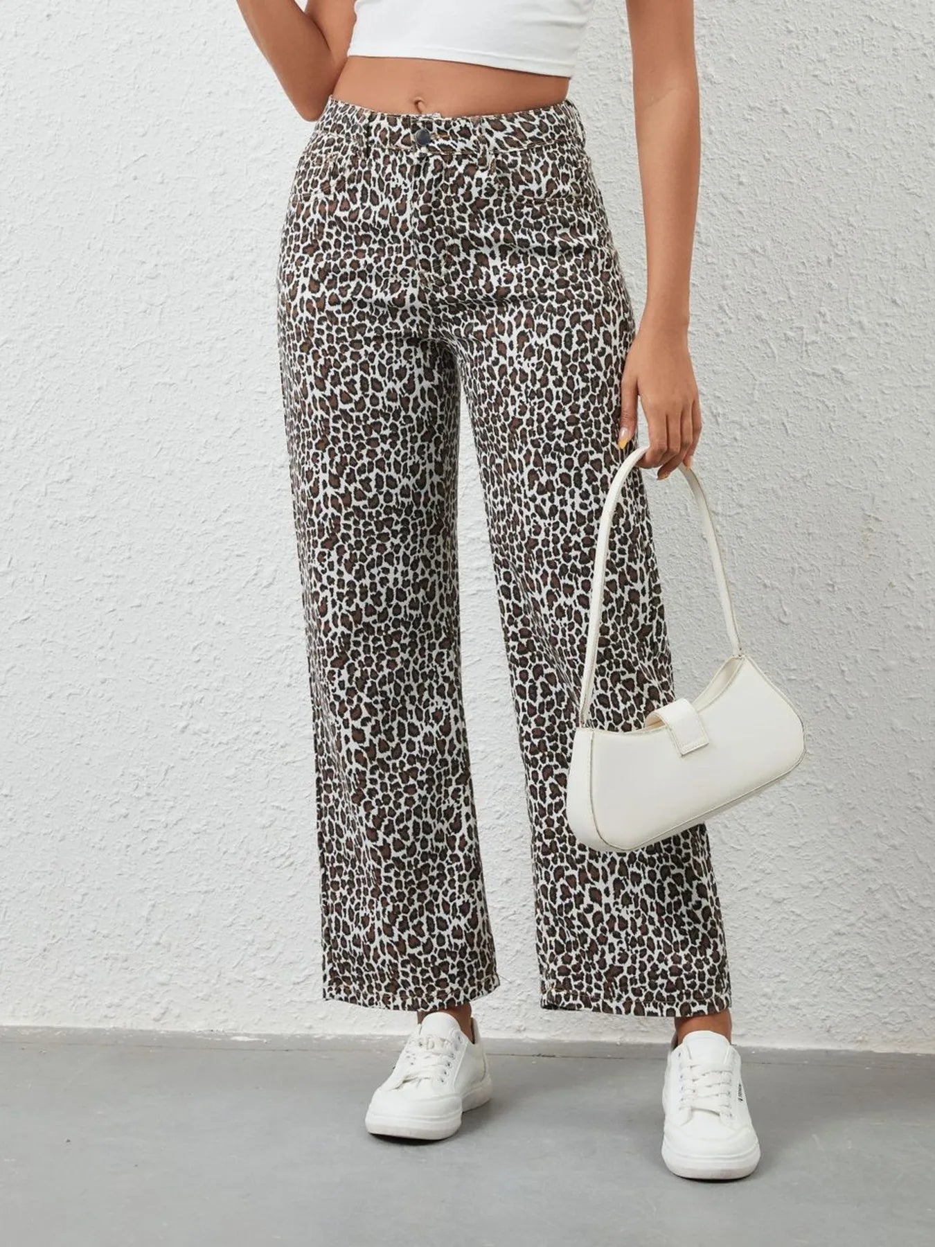 Person wearing leopard print pants and white sneakers holding a white handbag against a plain background