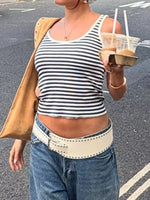 Person wearing a striped tank top and blue jeans, holding a drink, on a street.
