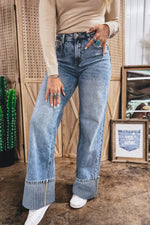 Person wearing blue jeans and a beige sweater in an indoor setting with a cactus and framed picture in the background.