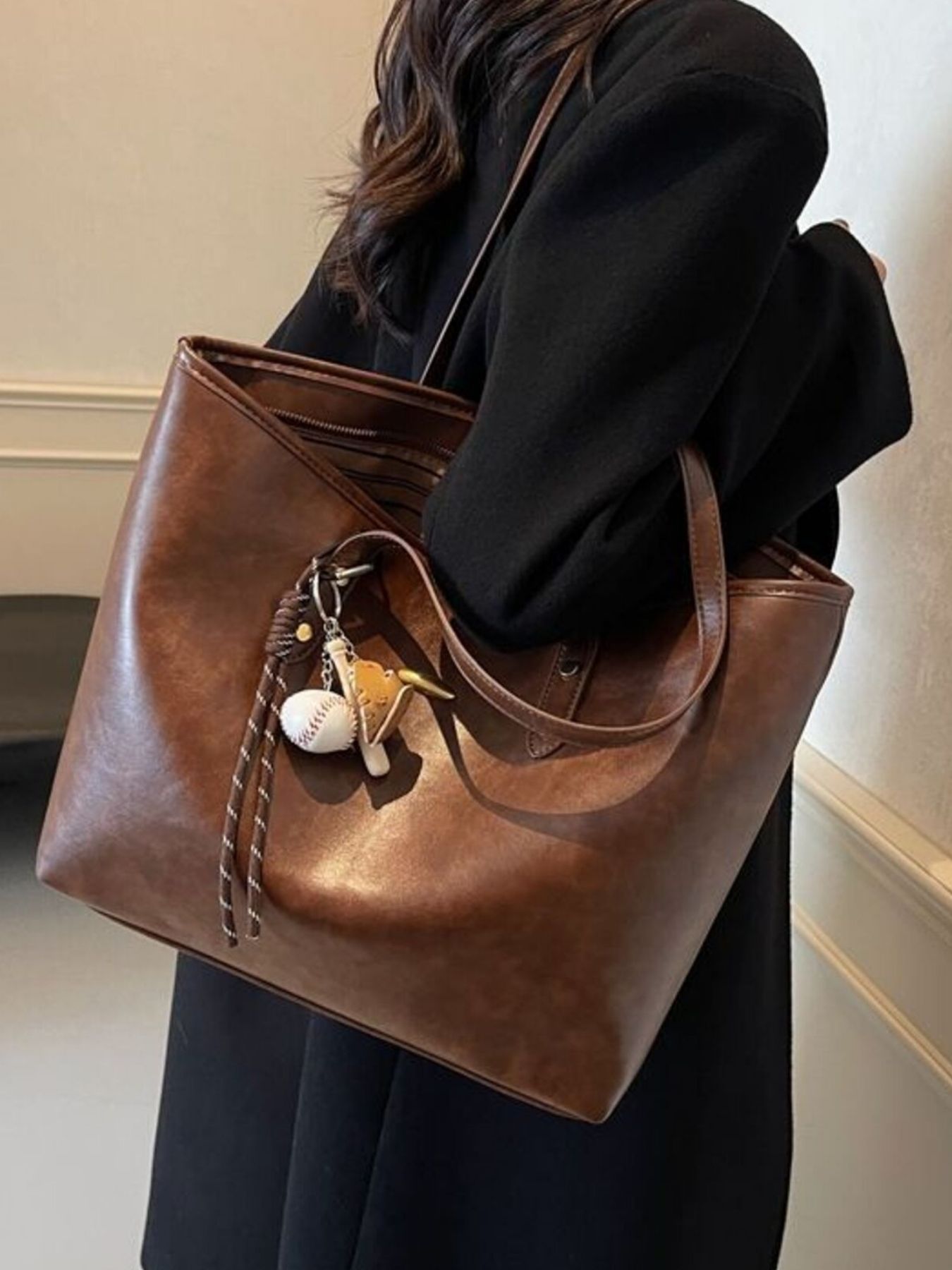 Brown leather tote bag with decorative keychain held by a person wearing a black coat.