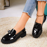 Black patent leather loafers worn with blue jeans on a textured surface.