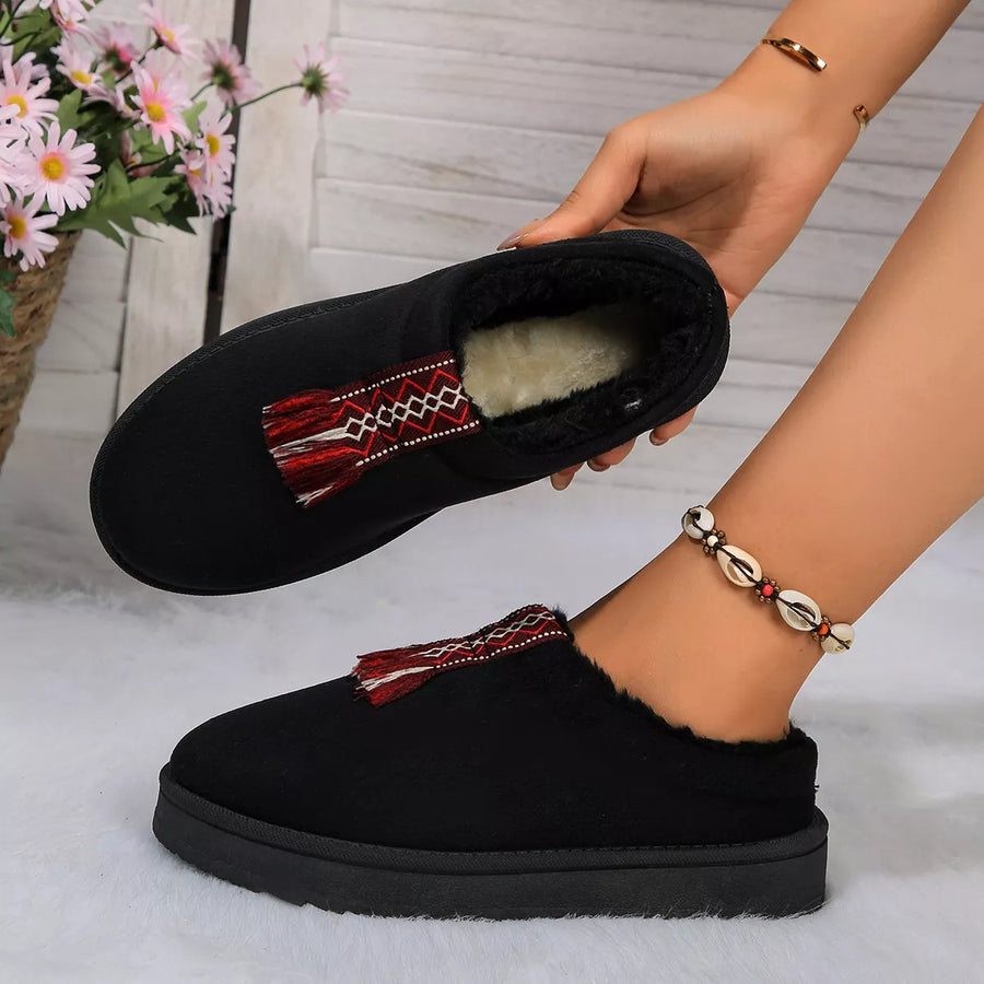 Black slip-on shoes with tassel details held by a person, with a blurred background of flowers and light-colored floor.