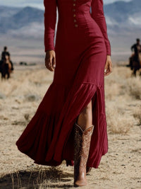 Person wearing a long burgundy dress and brown cowboy boots in a desert setting with mountains in the background.
