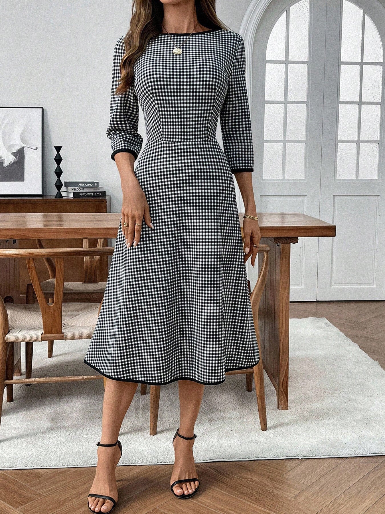 Woman wearing a black and white checkered dress in a room with wooden furniture and a large window.