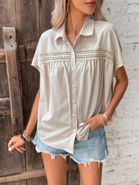 Woman wearing a beige button-up shirt with lace details and denim shorts against a rustic wooden and brick wall.