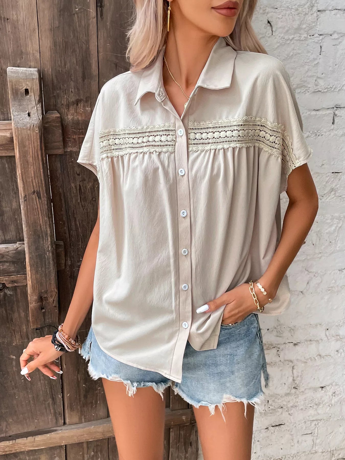 Woman wearing a beige button-up shirt with lace details and denim shorts against a rustic wooden and brick wall.
