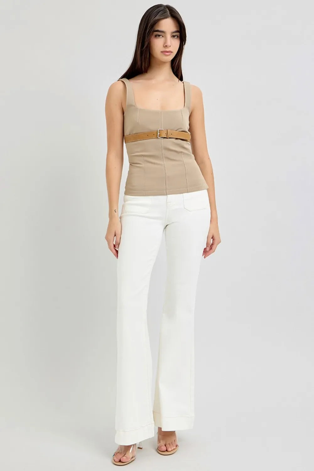 Woman wearing a beige tank top with a belt and white pants on a white background
