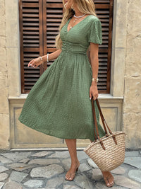 Woman in a green dress holding a straw bag against a stone wall.