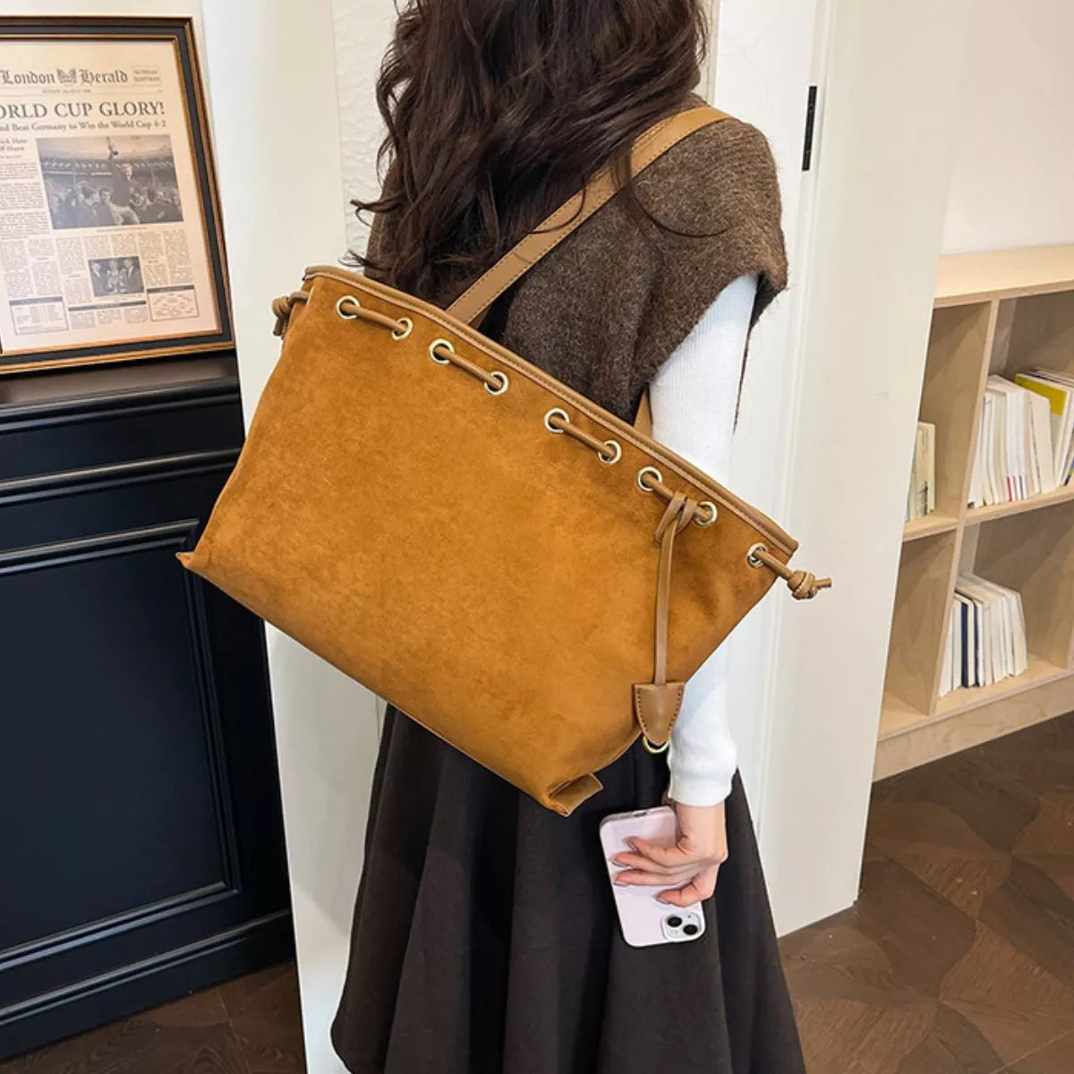 A woman wearing a brown sweater vest and black skirt carries a large mustard yellow suede shoulder bag with metal grommets in an indoor setting.