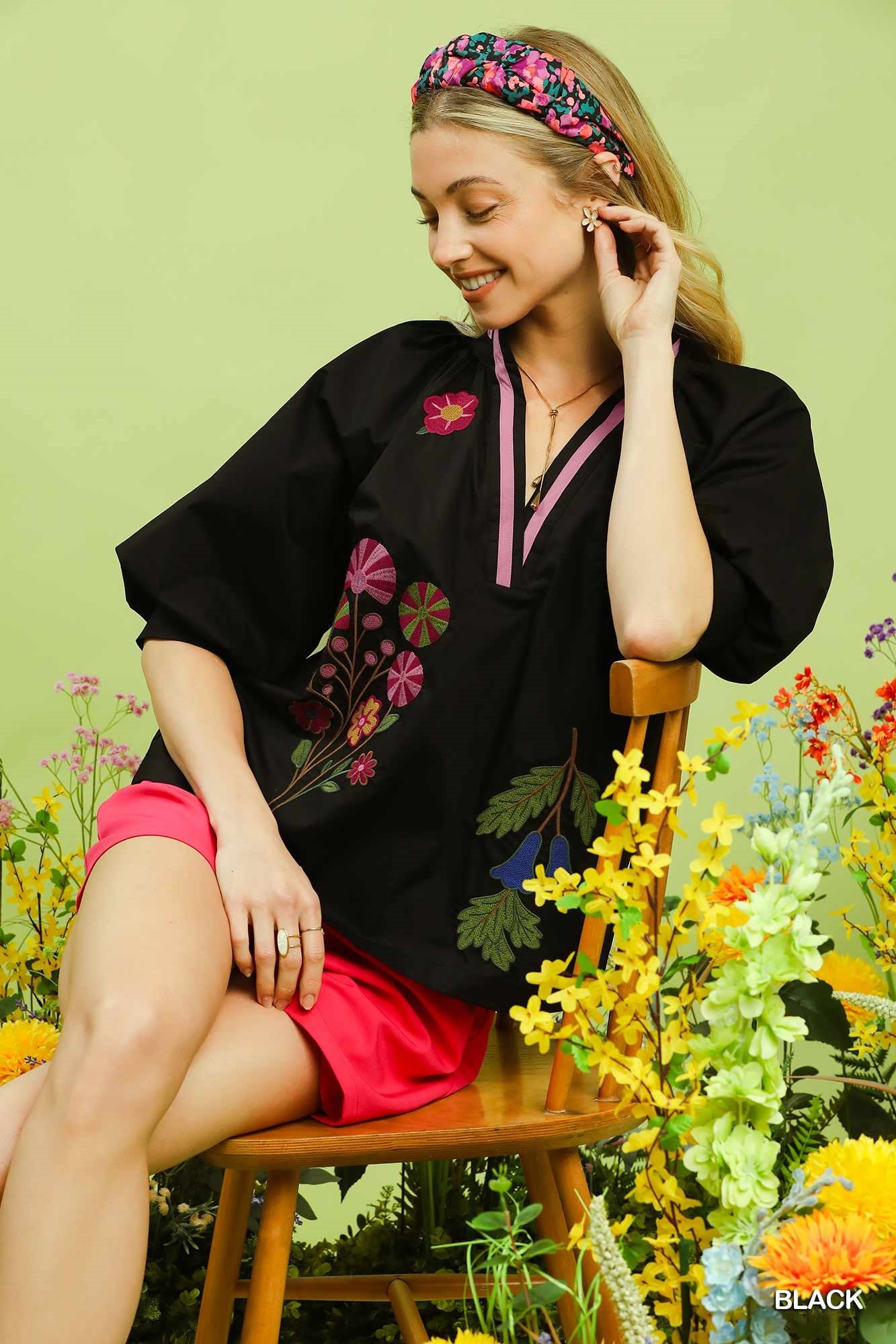 Woman sitting on a chair surrounded by flowers wearing a black embroidered top and pink shorts.