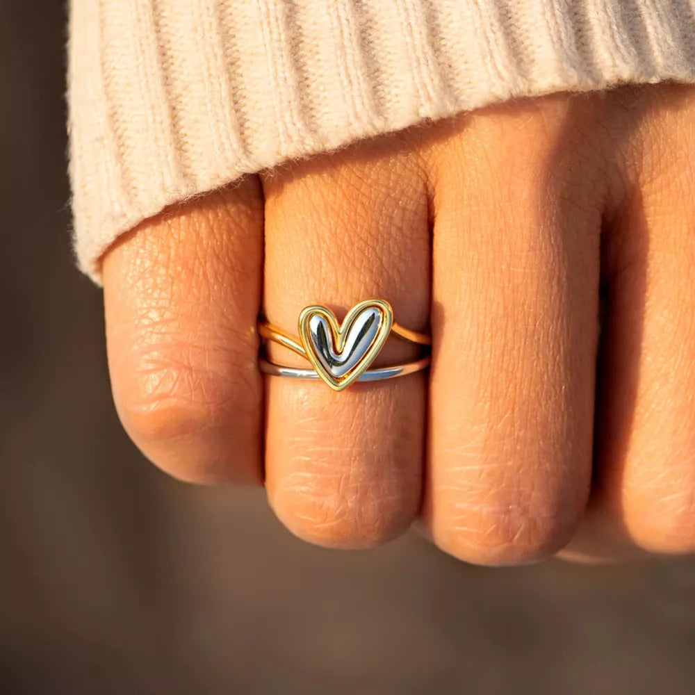 Hand wearing a gold and silver heart-shaped ring on a blurred background