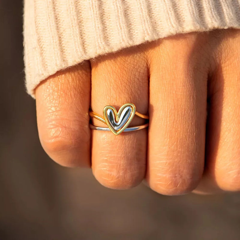Hand wearing a gold and silver heart-shaped ring on a blurred background