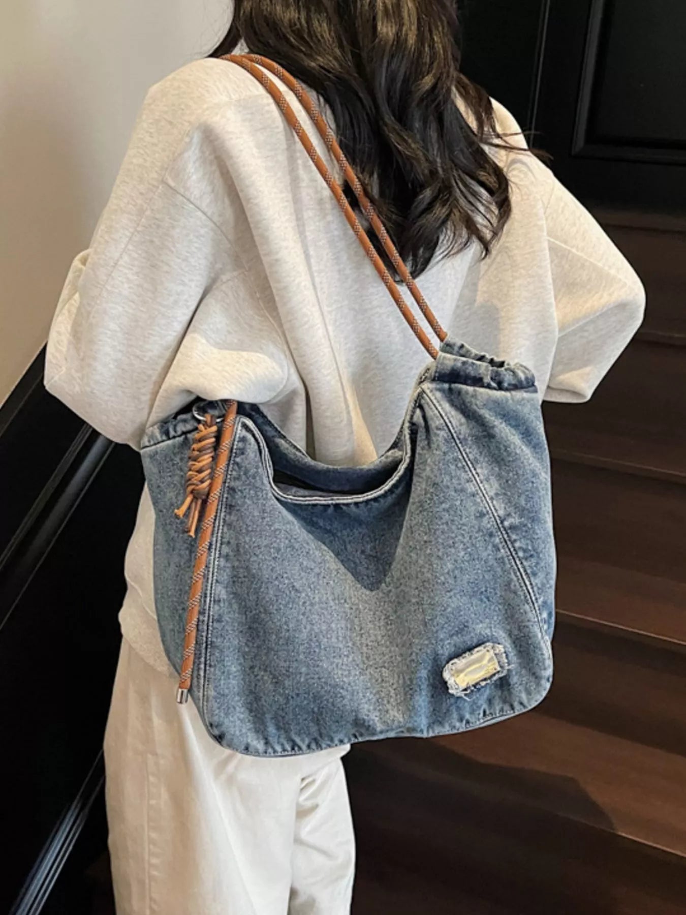 Person holding a denim handbag with brown straps indoors.