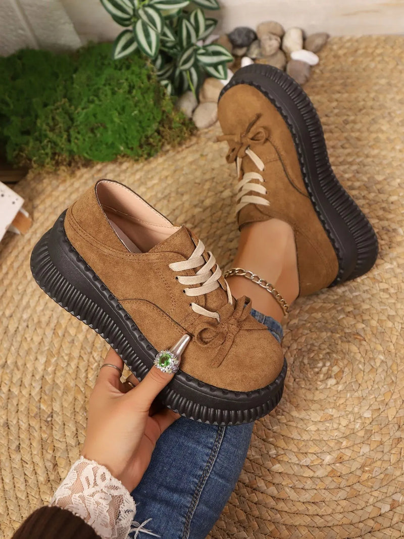 Brown suede shoes with black soles worn by a person on a textured surface.
